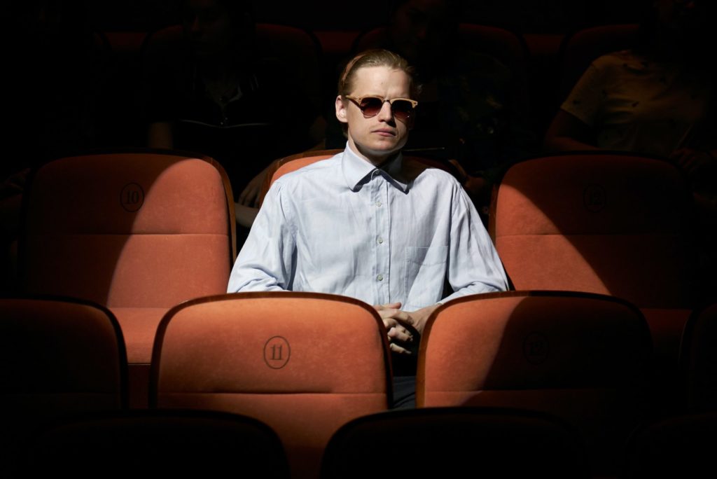 A man in dark glasses sits under a spotlight in the theatre seats